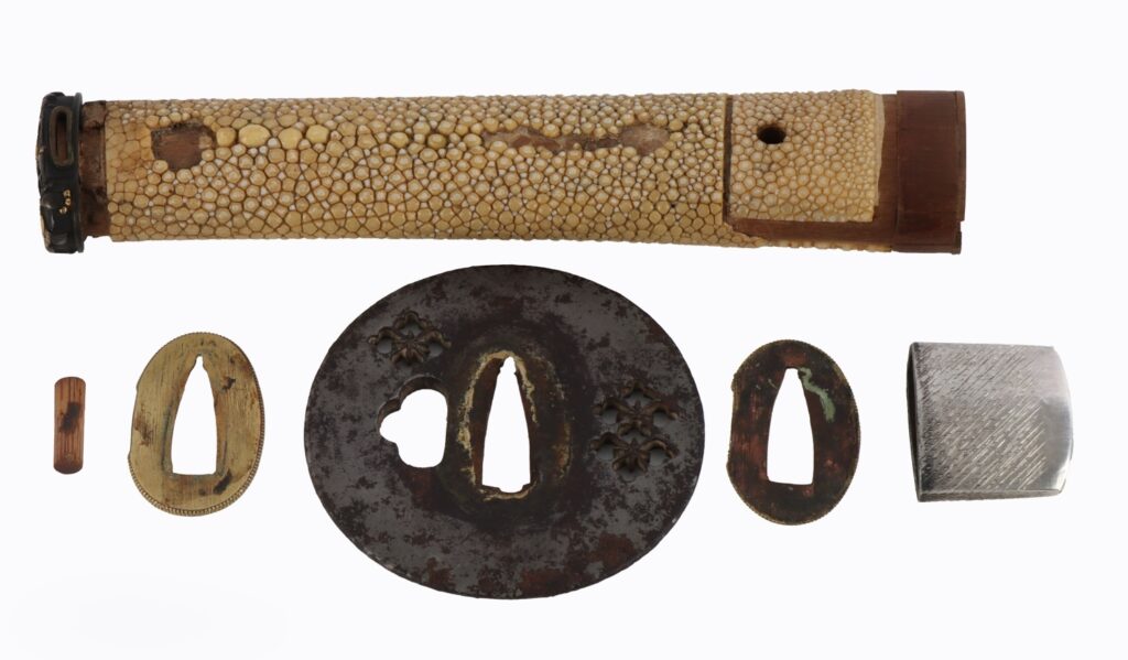 An arrangement of traditional Japanese sword fittings, including a tsuka (handle) wrapped in stingray skin, several tsuba (hand guards) in various materials, and other decorative elements.